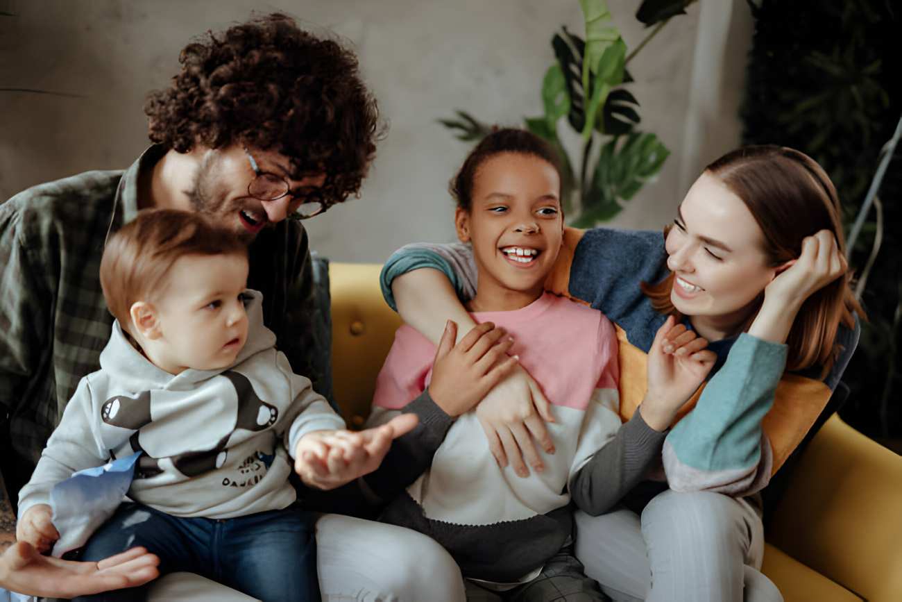 Happy family with Mom, Dad, and two young kids sitting on the couch, laughing and having fun together